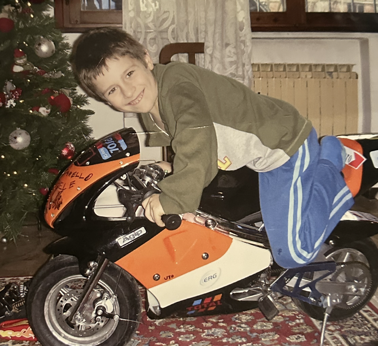 A five-year-old child on an orange racing pocket-bike beside a Christmas tree in Padova, Italy.